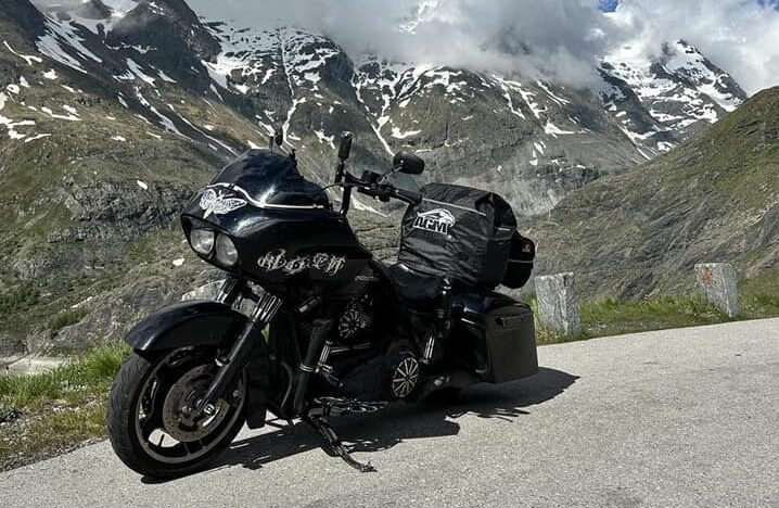 Harley-Davidson — Czarna Ćma — zaparkowany na alpejskiej przełęczy, w sercu gór ośnieżonych jeszcze końcówką zimy. Potężny motocykl kontrastuje z surowym krajobrazem — czarna sylwetka wpatrzona w niebo rozdzierane przez dramatyczne chmury. Samotna dusza w podróży, która nie zna celu — tylko kierunek.